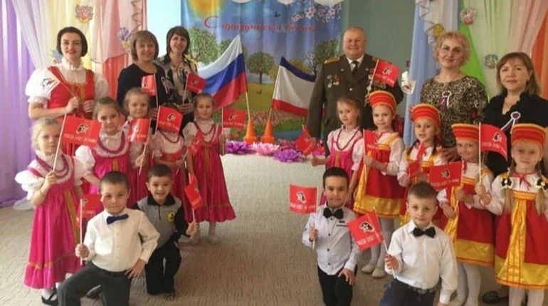 Діти в російських народних костюмах з прапорами РФ та військовим на святковому заході в дитячому садку на окупованій території.